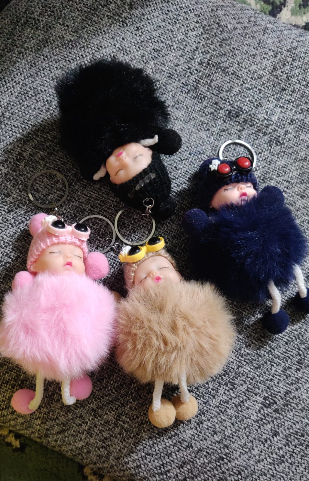 Four sleeping baby doll pom pom keychains in pink, beige, black and navy on grey fabric